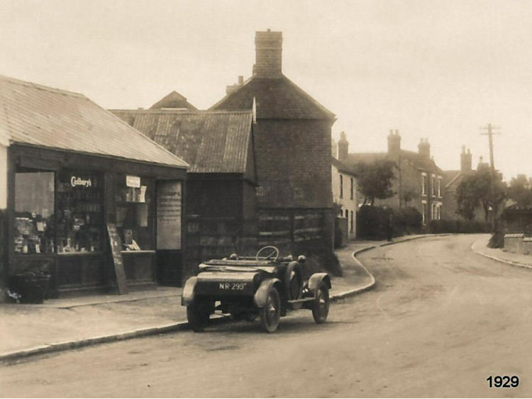 Shop and buildings along the road with a motor car, dated 1929.